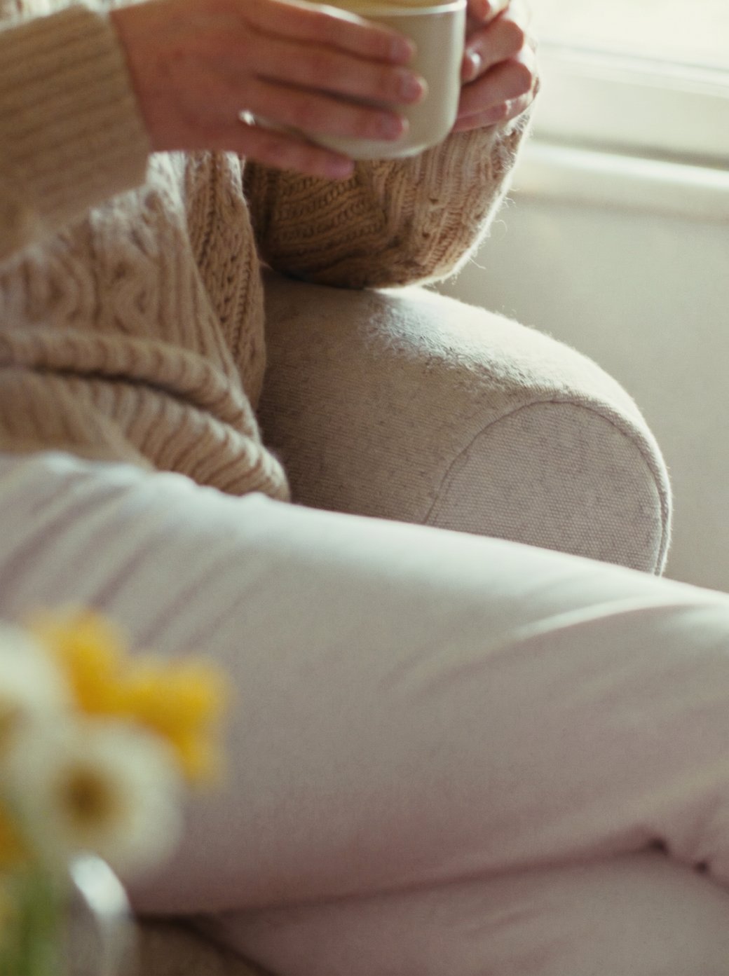 Eine Frau im warmen Strickpullover hält in einem Sessel eine Tasse Tee, ein ruhiger Moment für sich